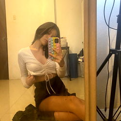 Ariel little, posing in a mirror with her hands on hips and wearing cropped white top and skirt, in what appears to be an indoor setting.