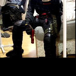 An image of a person dressed in tactical gear, holding a device in one hand and sitting on the floor against a white wall. The individual appears to be a young male, possibly a costume or cosplay event given the attire.
