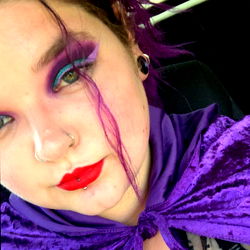 A person with vibrant makeup and purple hair, wearing a cape and glasses