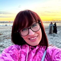 BustyBexx, a California-based model and influencer, poses for a selfie on the beach at sunset. She is wearing glasses, a pink hoodie, and has vibrant red hair.
