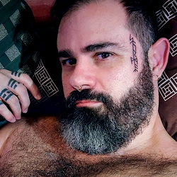 Storm Sol, a bearded and tattooed man showcasing his muscular physique in a bedroom setting.