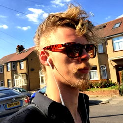 A young man, Arthur, strolling down a street in the United Kingdom. Wearing sunglasses and sporting a beard.