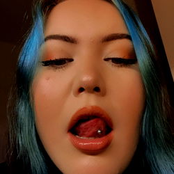 Amelia, a 25-year-old model from the UK, poses playfully with her tongue out. Her blue hair and striking makeup add a vibrant touch to her look.