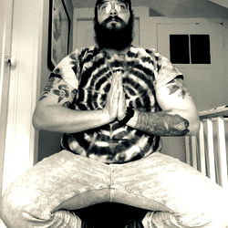 Bearded Man in Tie-Dye Shirt and Jeans Sitting on Floor with Hands Together