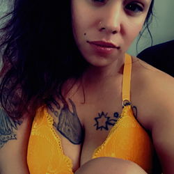 Mandi Mae, a young woman posing in a vibrant yellow bra and black tattoos against a gray wall,